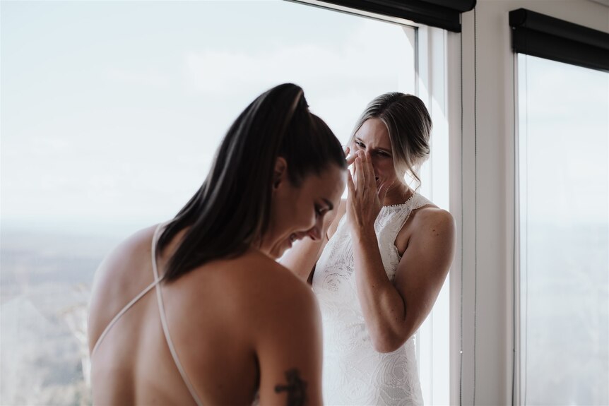 A bride tears up as she looks at her wife in her wedding dress.