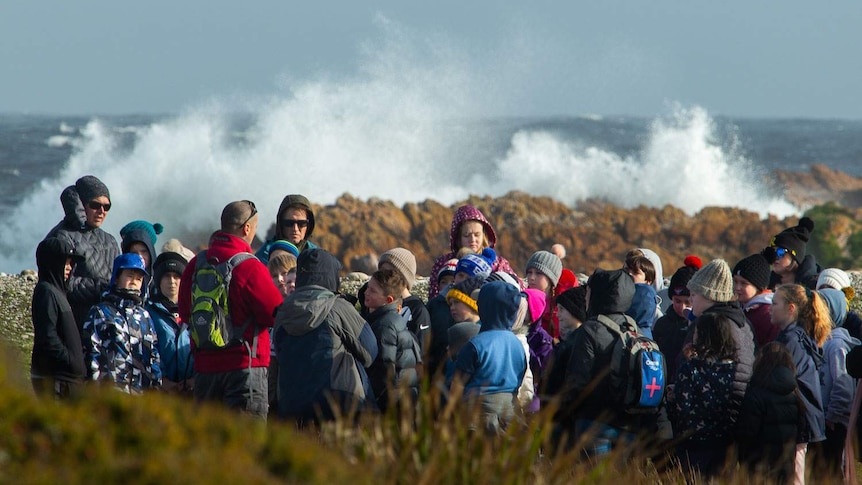Schoolkids wear beanies and raincoats and listen to a guide as a big waves explode on the coastal rocks behind them