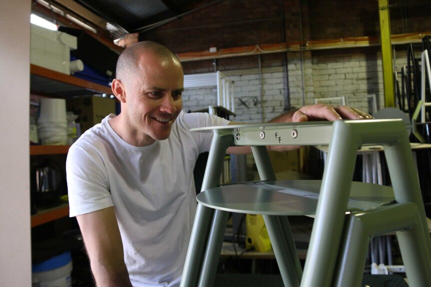 Elliot works on a piece of furniture in his workshop, smiling.