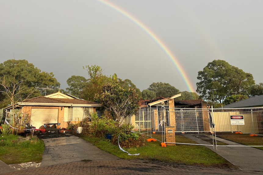 Two neighbouring houses with a rainbow seen in the background. Both properties have fire damage, with a fence surrouding one.