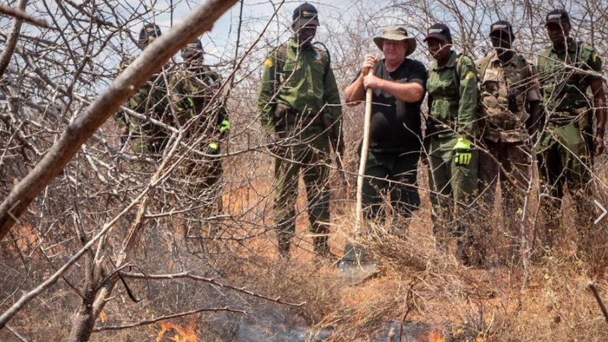 A group of men stand in the bush and watch on as a small fire burns.