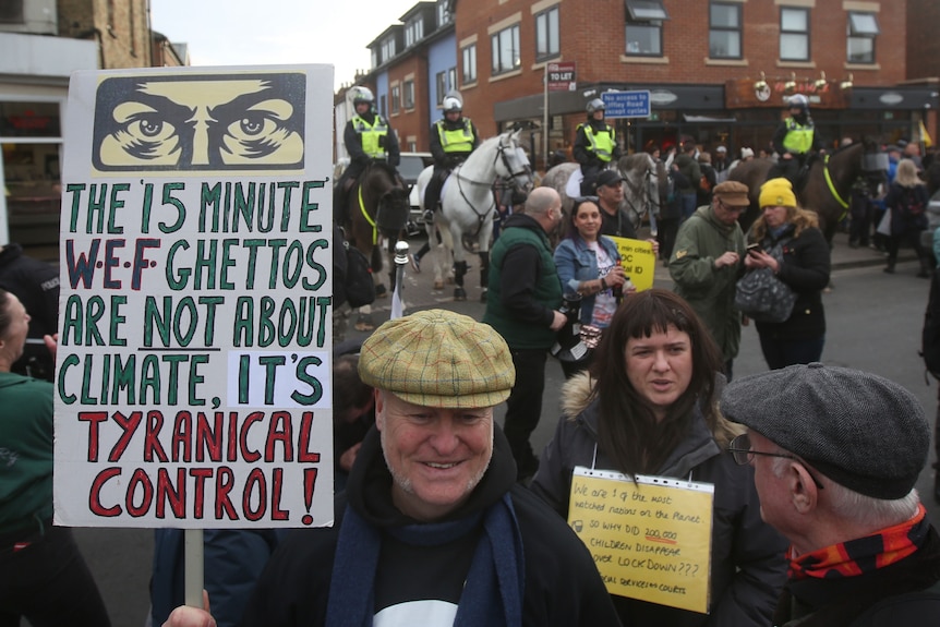A man at a protest with a sign reading 'The 15 minute WEF ghettos are not about climate. It's tyrannical control'