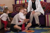 Ms Waller sits on a rug and claps her hands and looks at her 14-month-old daughter, who is listening to a story being read..