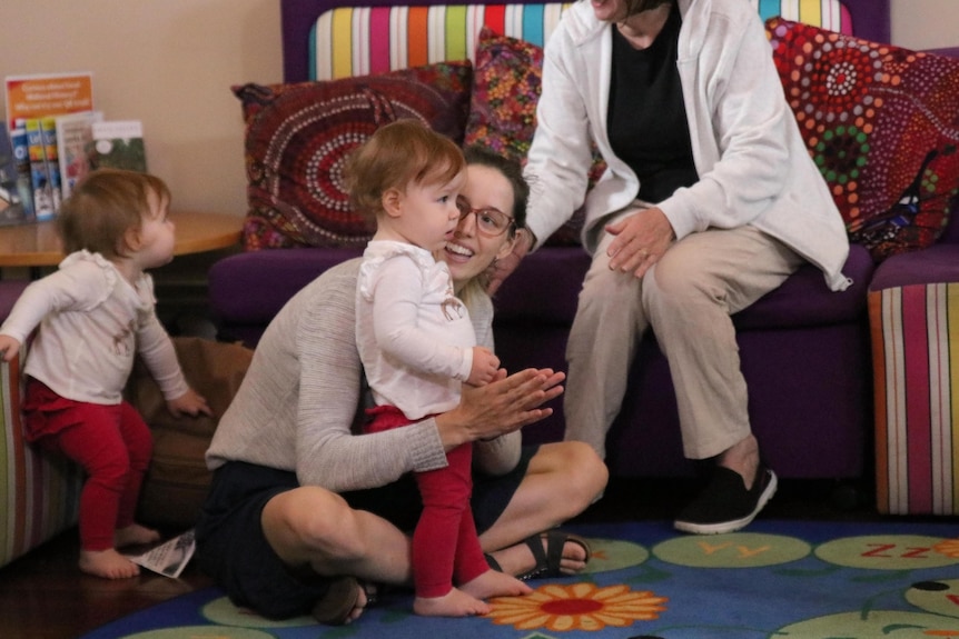 Ms Waller sits on a rug and claps her hands and looks at her 14-month-old daughter, who is listening to a story being read..