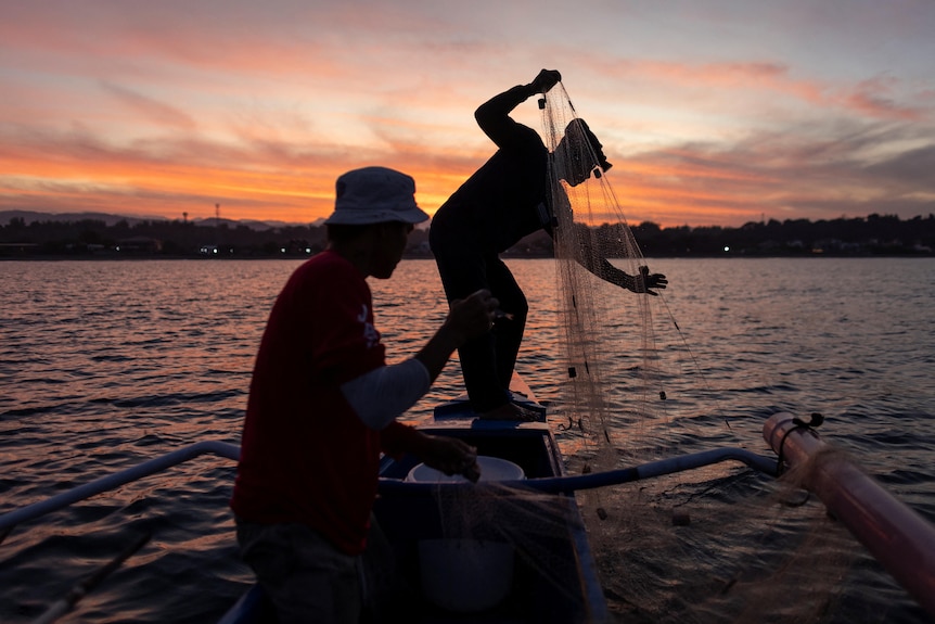 A person pulls a net from the water as another looks on. 
