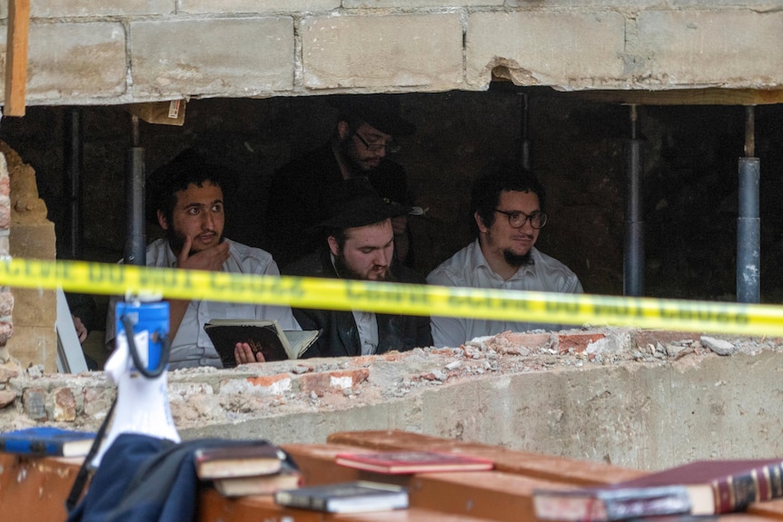 A group of male Jewish students sit behind an opening in a wall. Police tape and religious books on desks in foreground