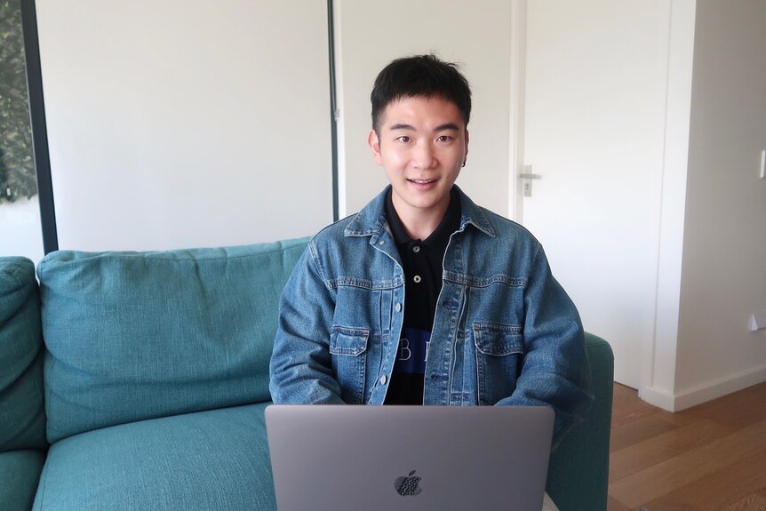 A young Chinese man sitting at a computer and smiling in a denim jacket. 