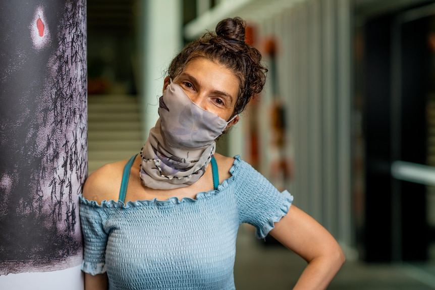 A woman with a scarf over her face looks at the camera with a cocked head. She has brown hair and is wearing a blue shirt.