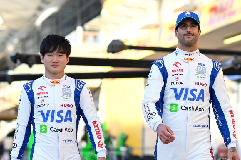 Yuki Tsunoda and Daniel Ricciardo of Visa Cash App RB F1 team walk side by side down pitlane in full racing suits.