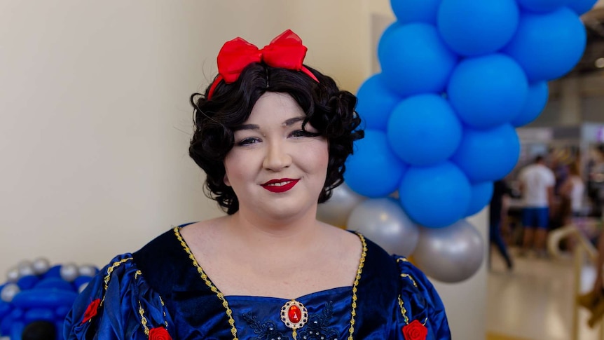 A woman wears black wig with red bow on top, puffy sleeves with flowers. White make-up and red lips. Blue balloons in background
