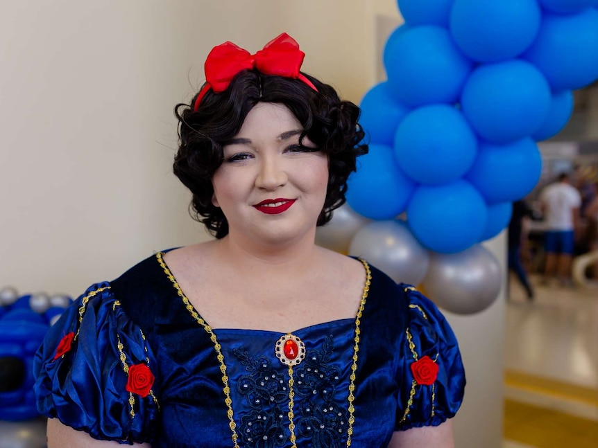 A woman wears black wig with red bow on top, puffy sleeves with flowers. White make-up and red lips. Blue balloons in background