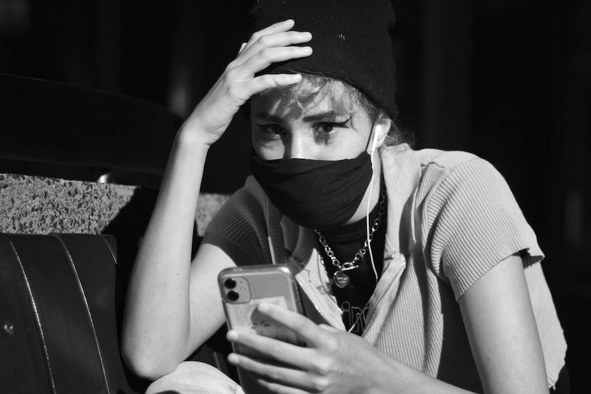 A young woman sits on a bench wearing a face mask. She has one hand on her head and the other holding her mobile phone.