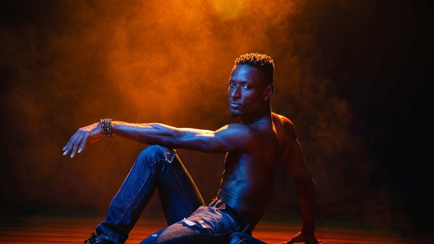 A dramatic colour wide photo of Dashaun Wesley in a dance pose on the floor in front of dark background.