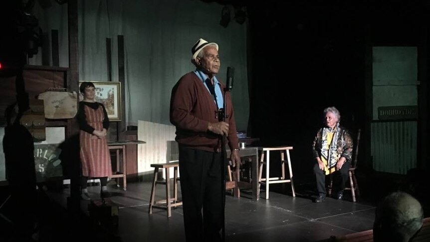 Older man on stage speaking into microphone with two women standing at the back of the stage