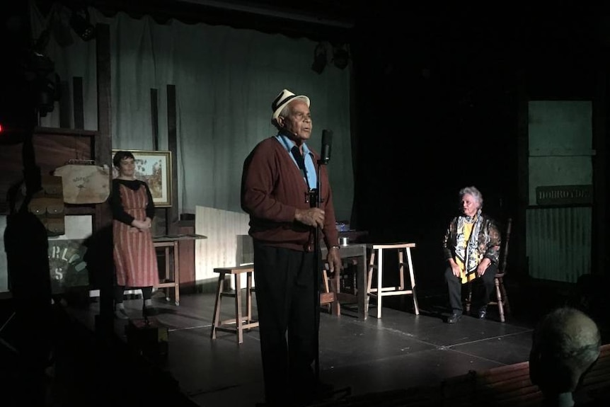 Older man on stage speaking into microphone with two women standing at the back of the stage.