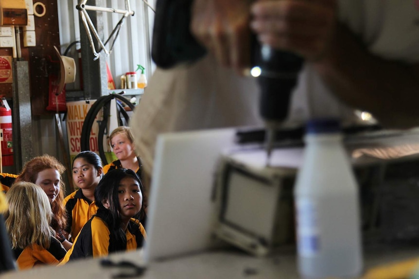 A close-up photo of a drill being applied to a row of plastic cubes, with schoolchildren in focus in the background.