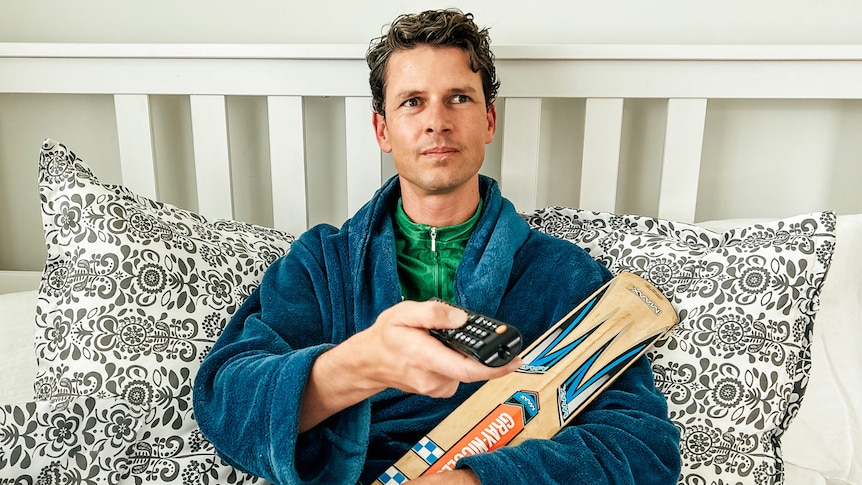 a man sits in bed in a robe and cycling shirt with a remote and cricket bat in hand