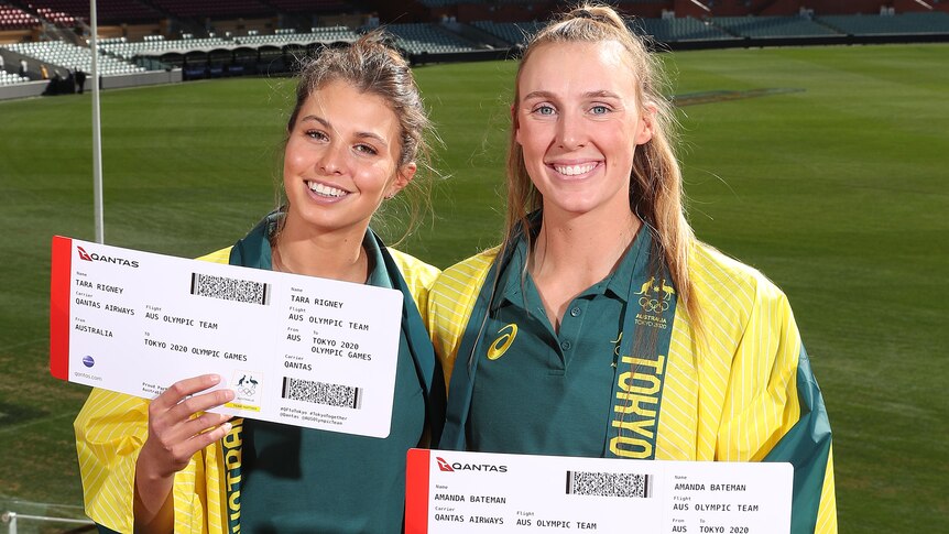 Two women holding large boarding boarding passes and wearing green and gold robes over green uniform shirts.
