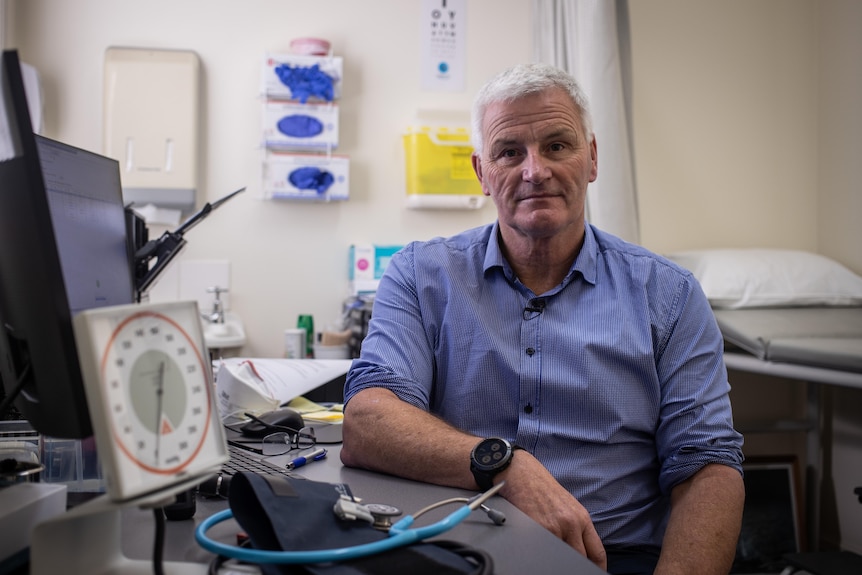 A man leans against his desk, with medical equipment in the backgrounbd and foreground.