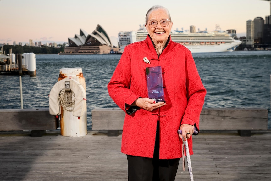 A woman in a red jacket holds an award in front of the Sydney Harbour. 
