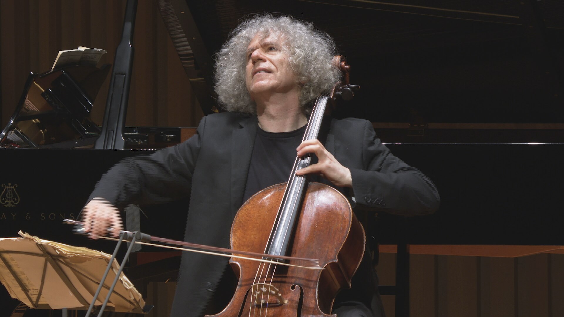 Dressed in 'concert blacks' a long-grey-haired cellist plays in front of a piano.