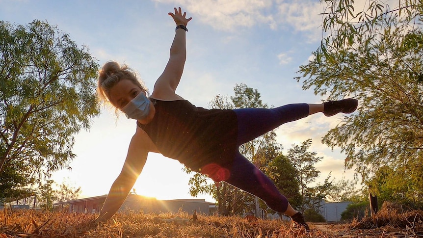 Low shot in bush into the sunrise with lady standing on one hand and one foot wearing surgical mask