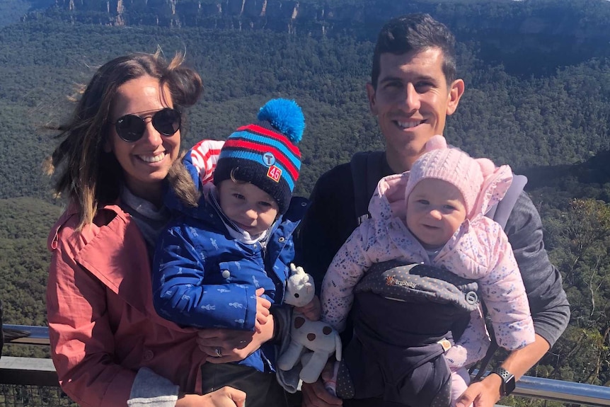 A man and a woman stand together smiling at a bush lookout location, each holding a child in their arms.