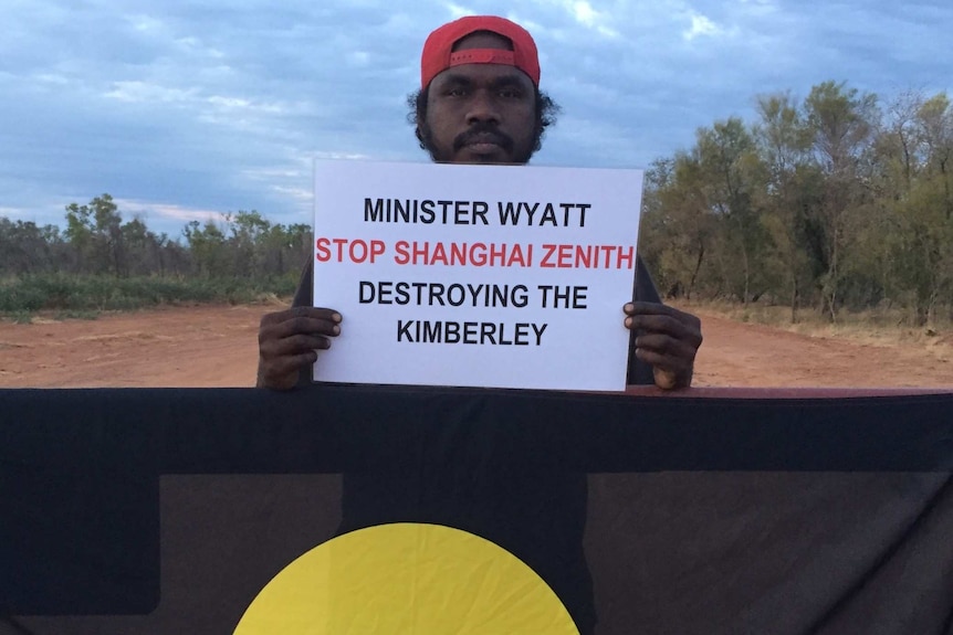 A man holds a sign up in behind an Aboriginal flag in front of a red dirt road
