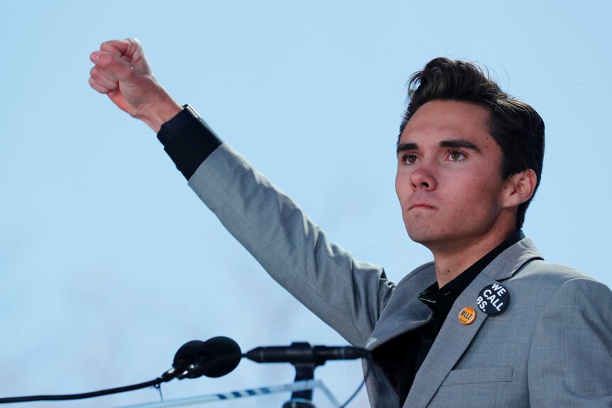 David Hogg purses his lips as he thrusts his fist into the air while standing behind a microphone.