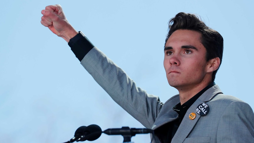 David Hogg purses his lips as he thrusts his fist into the air while standing behind a microphone.