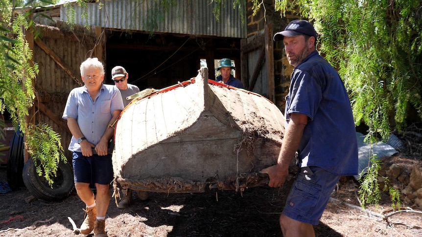 Four men carry an upturned old wooden boat