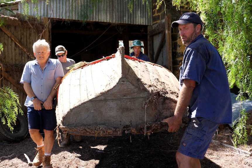 Four men carry an upturned old wooden boat