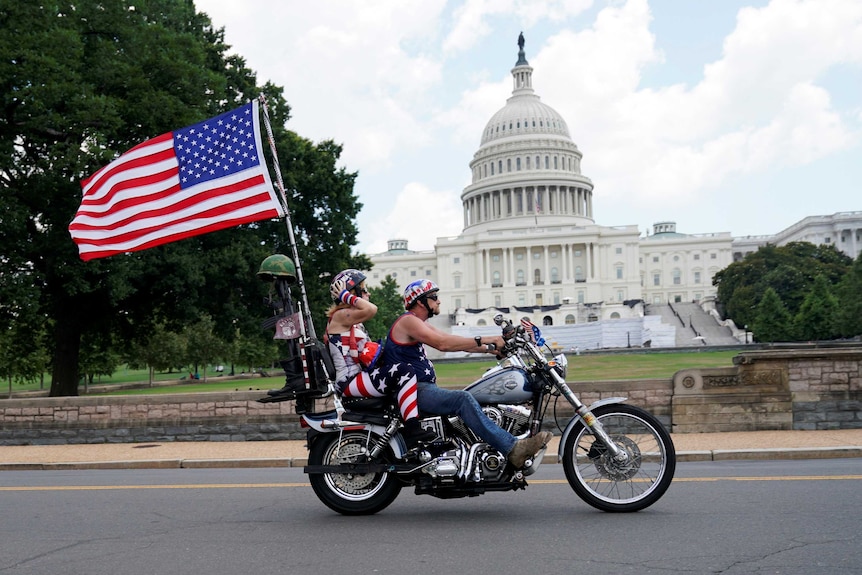 A middle-aged couple ride a big motorbike and fly a US flag as they ride past a white building.