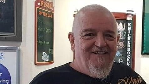 An older, heavyset man stands smiling in what appears to be a bistro.