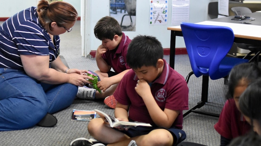 Students and a parent sit on the floor of a classroom, reading books.