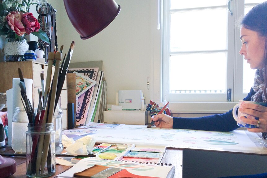 Lisa Schroder holds a coffee in one hand as she paints with the other in her studio.