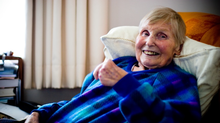 A older woman sitting with a cup of tea in her dressing gown and smiling.