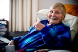 A older woman sitting with a cup of tea in her dressing gown and smiling.