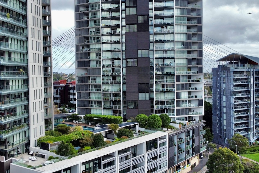 Four newly constructed apartment blocks surrounded by green trees and grass with grey clouds in the background.