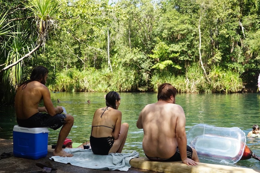 Three people sit looking out to the water at Berry Springs. A man on the left sits on an esky, there are trees on the other side
