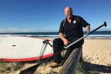 Dean McSporran sits at the beach with his paddle board and oar.