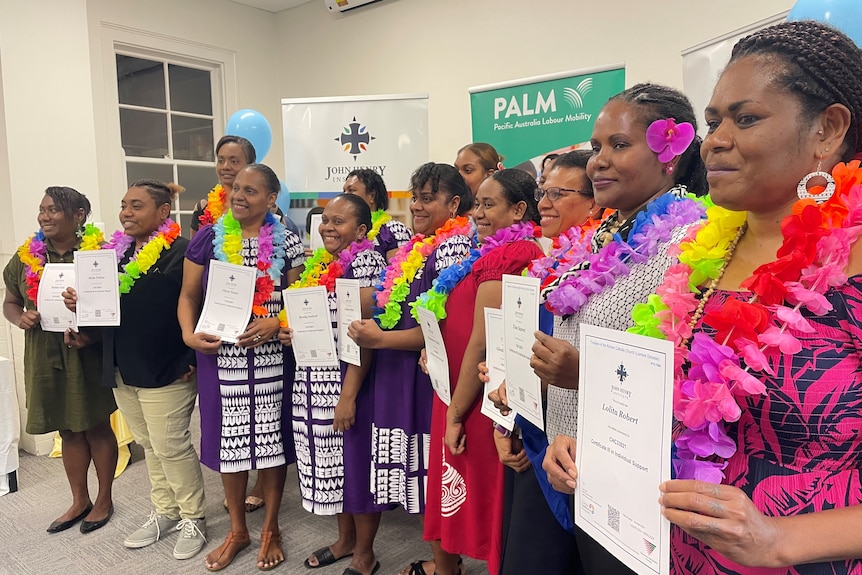 A group of women from Vanuatu, wearing tradional bright Pacific island dresses stand in a line holding graduation certificates.