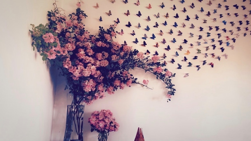 Butterflies made from paper cover a white wall and blend into a vase of pink flowers.