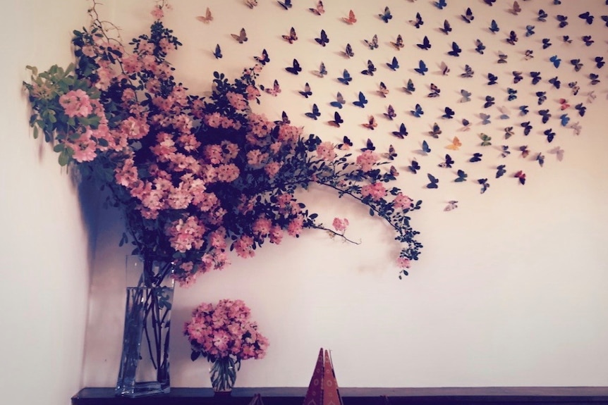 Butterflies made from paper cover a white wall and blend into a vase of pink flowers.