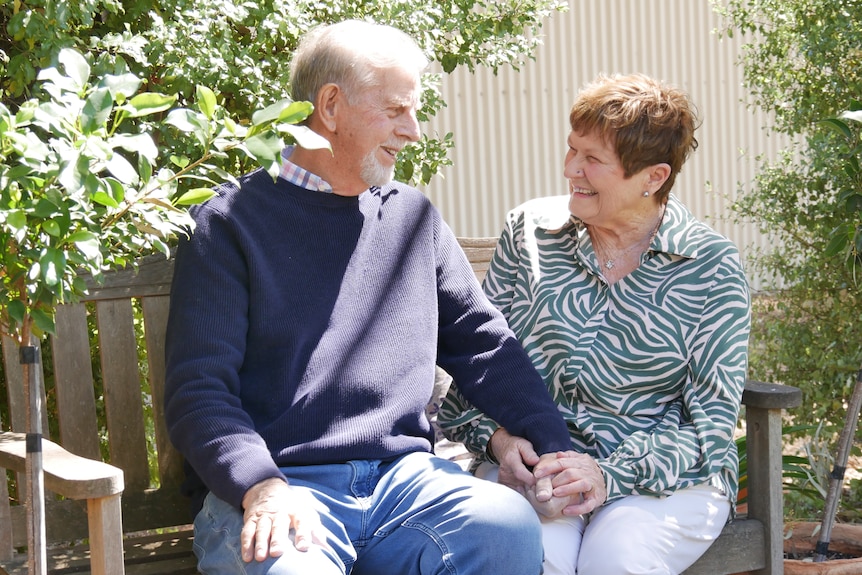 Bob and Jan Moulton sit on a bench at their home in Melrose, holding hands and smiling. 