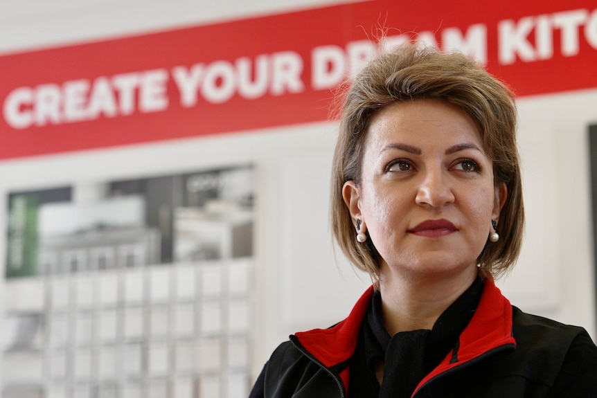 A woman with short brown hair tucked behind her ears wearing pearl earrings stands in a kitchen showroom looking to her left.