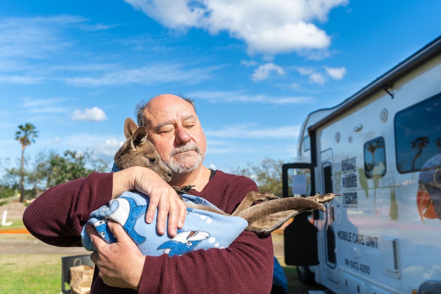A man closes his eyes as he stands next to a caravan beneath a blue sky, cuddling a baby kangaroo wrapped in a blanket.