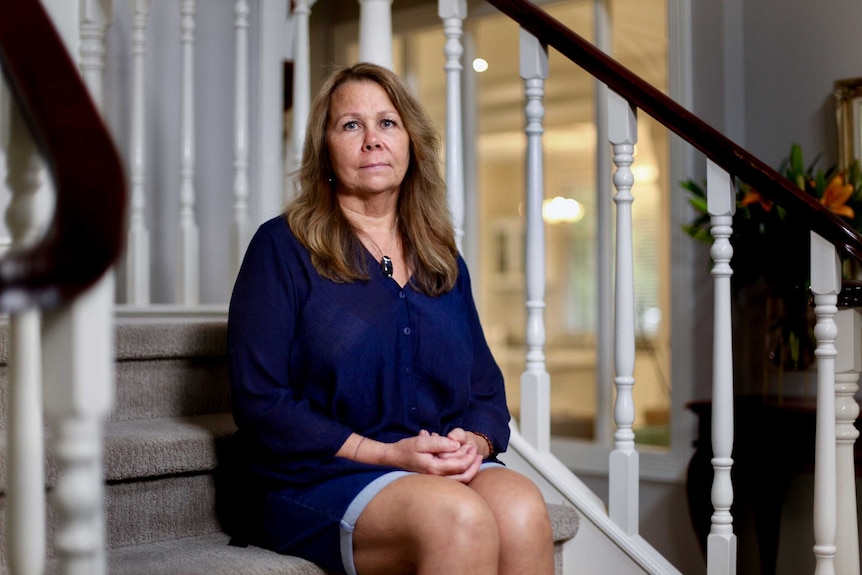 Woman with brown hair wearing blue blouse sits on stairs with polished wood banister and large room in background