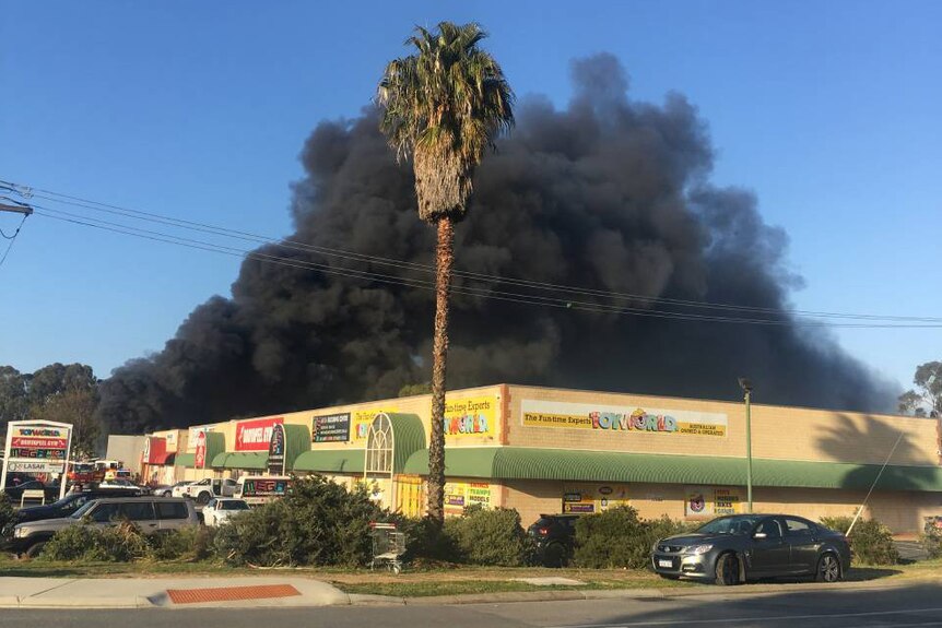 Thick black smoke billows into a blue sky above a retail building housing various shops.