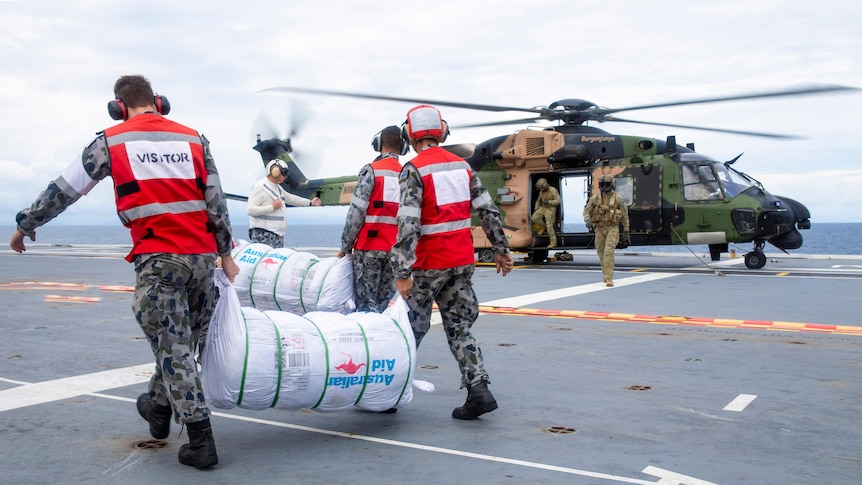 Defence personnel carry large bundles of aid towards a helicopter that's parked on a navy vessel.